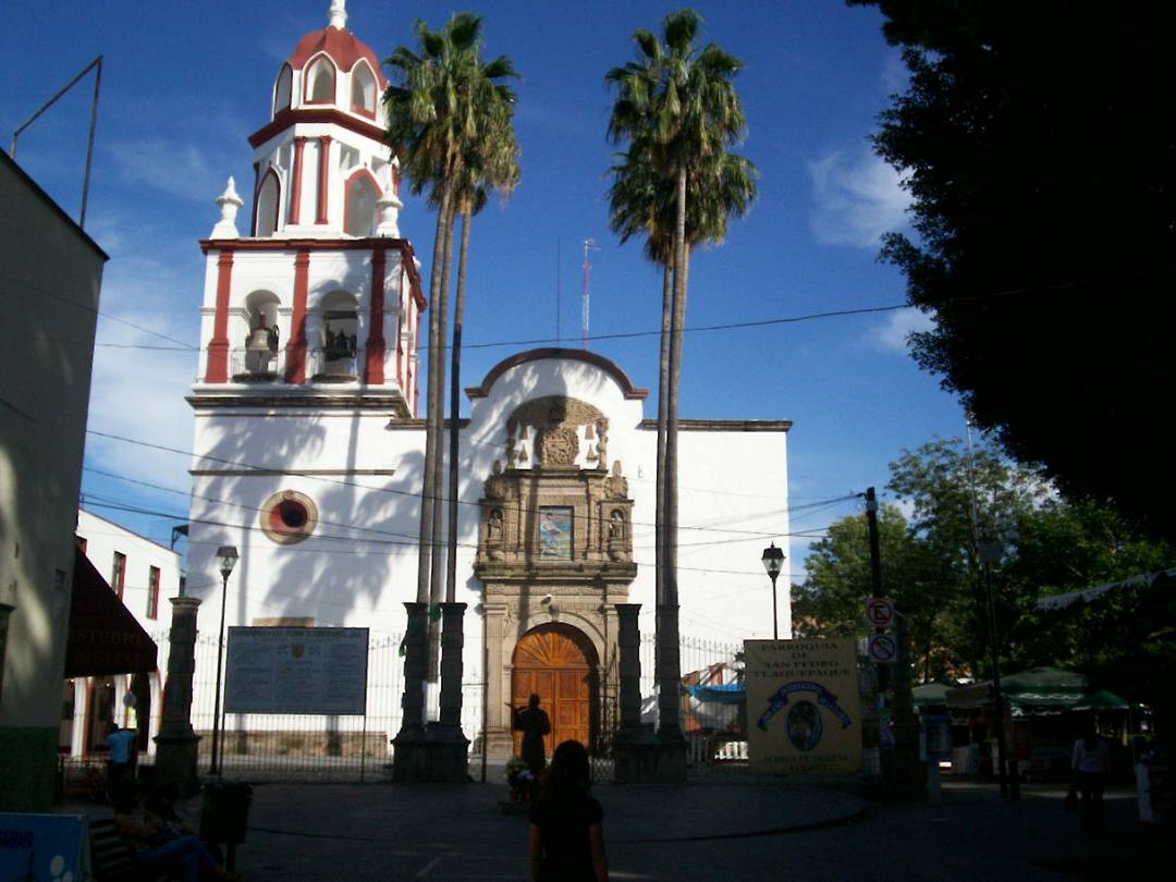 Iglesia con torre alta y cúpula roja, rodeada de palmeras y personas en la plaza.