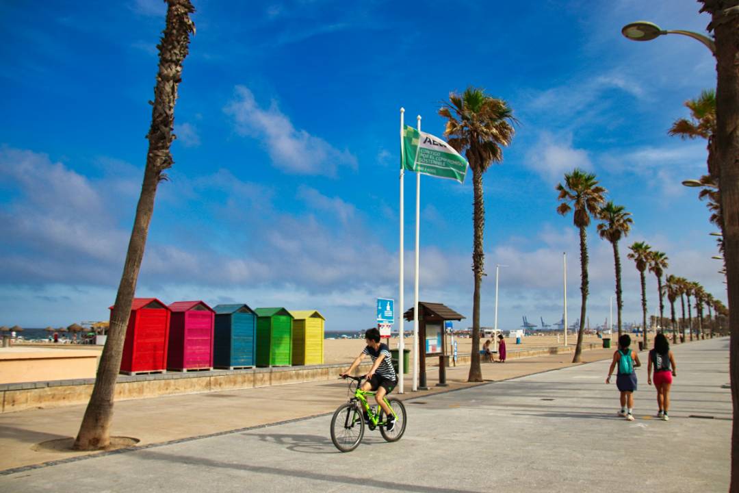 A sunny seafront promenade in Salou, Spain, features a cyclist riding past colorful beach huts, tall palm trees, and flagpoles, with pedestrians strolling along the wide pavement under a bright blue sky with wispy clouds.