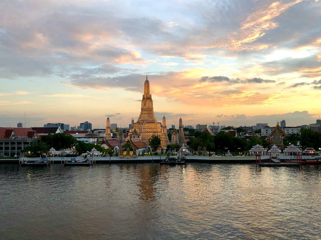 An ornate temple spire on a riverbank at sunset with a city skyline in the background.