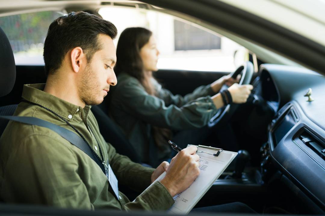 A woman drives a car while a man in the passenger seat writes on a clipboard.