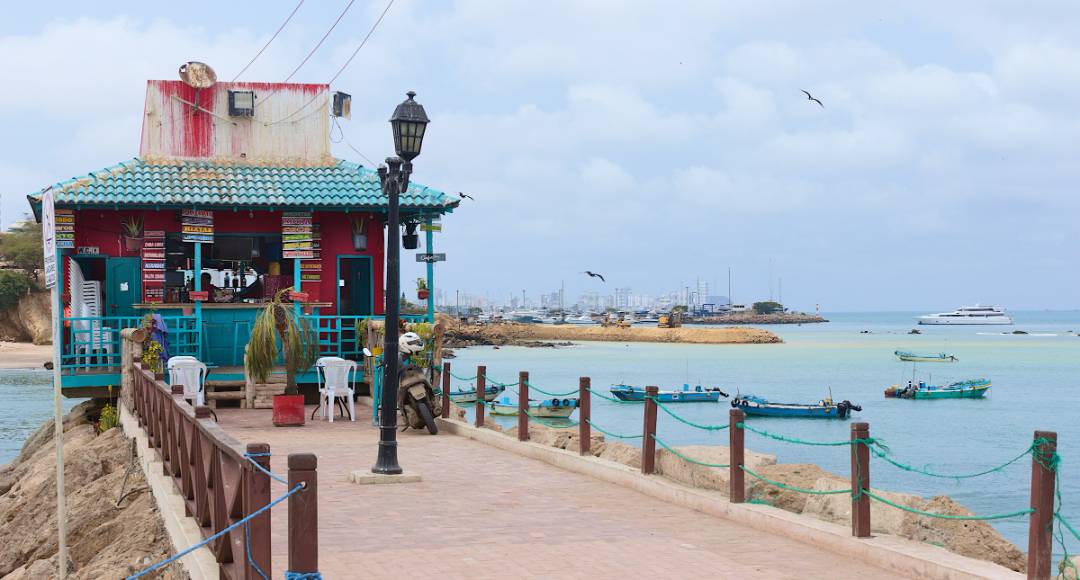 A vibrant beachside café with a turquoise roof sits along a coastal promenade in Ecuador, overlooking calm turquoise waters dotted with small fishing boats and a distant marina, under a partly cloudy sky with seabirds in flight.