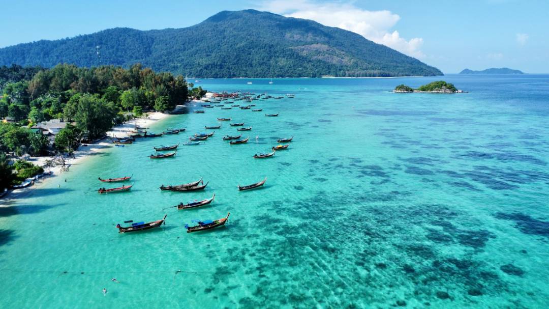 A picturesque tropical bay in Thailand features a sandy beach lined with traditional longtail boats, crystal‑clear turquoise waters revealing coral formations, lush green vegetation, and a forested mountain rising in the background under a bright blue sky with scattered clouds.
