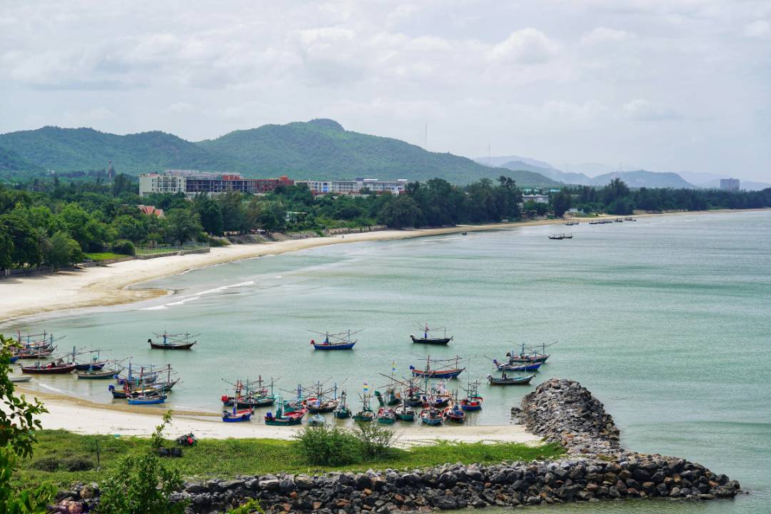A scenic coastal view in Thailand features a curving sandy beach lined with traditional fishing boats, backed by lush green hills and modern resort buildings under a soft, cloudy sky.