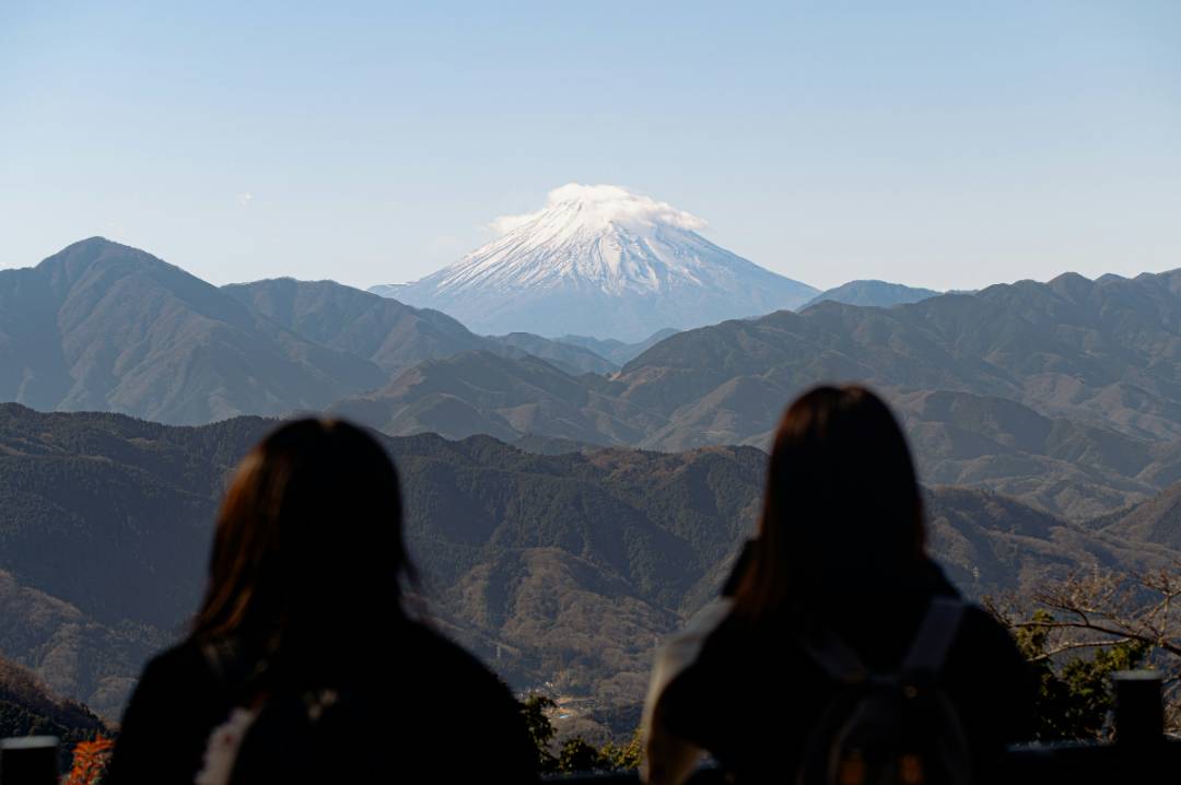 Two women admire the view of Mount Fuji from a mountain, surrounded by lush greenery and a clear blue sky.
